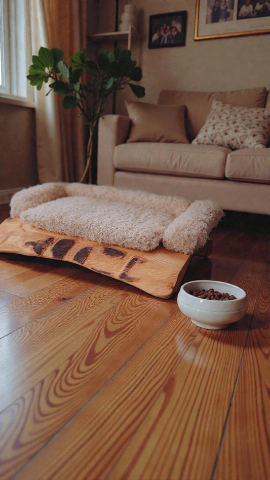 Solid oak pet bed on a wooden floor with a bowl of food nearby in a living room setting.